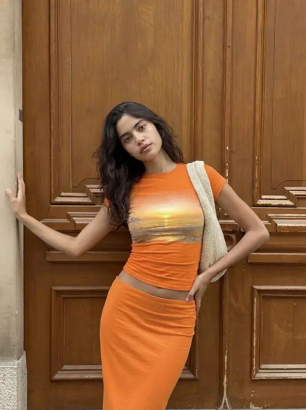 AI actor Priya in an orange sunset top and skirt by a wooden door, from Pixraft Marketing Studio